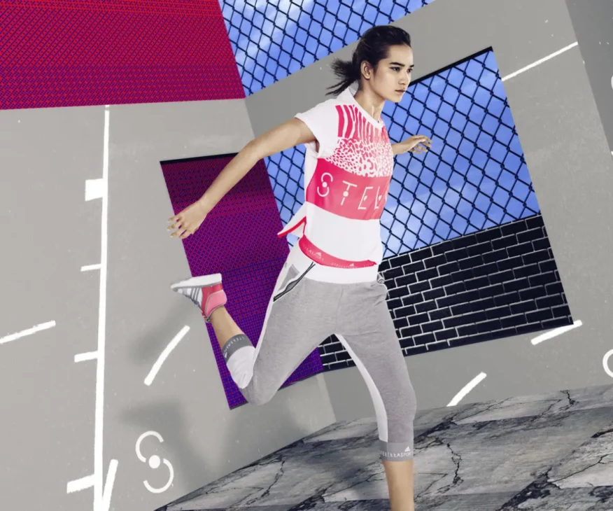 Model leaping in white graphic Adidas StellaSport logo tee and grey pedal pushers