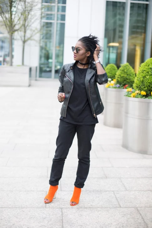 Full-length street style outfit showing black high-waisted jeans, biker jacket, and bright orange heels against London greenery
