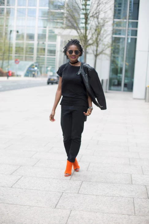 Full-length street style shot of an all-black outfit with leather jacket and bright orange heels walking outdoors