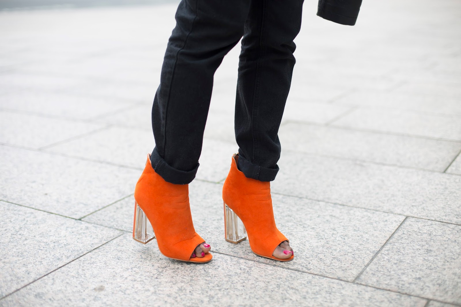 Detail shot of bright orange heels contrasting against high-waisted black slim jeans on urban pavement